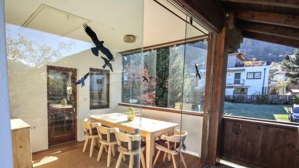 Een moderne kamer met een houten eettafel en stoelen, grote ramen met uitzicht op een tuin en gebouwen, en een plafond met silhouet van een vogel.