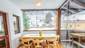 Een eetruimte met een houten tafel en stoelen, een gele vaas en uitzicht op huizen door grote ramen.