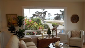 A living room with a sofa, armchair, and a glass coffee table with candles. There is a window with a view of a road and greenery outside. - Perfect Pink Paradise (Cayucos)