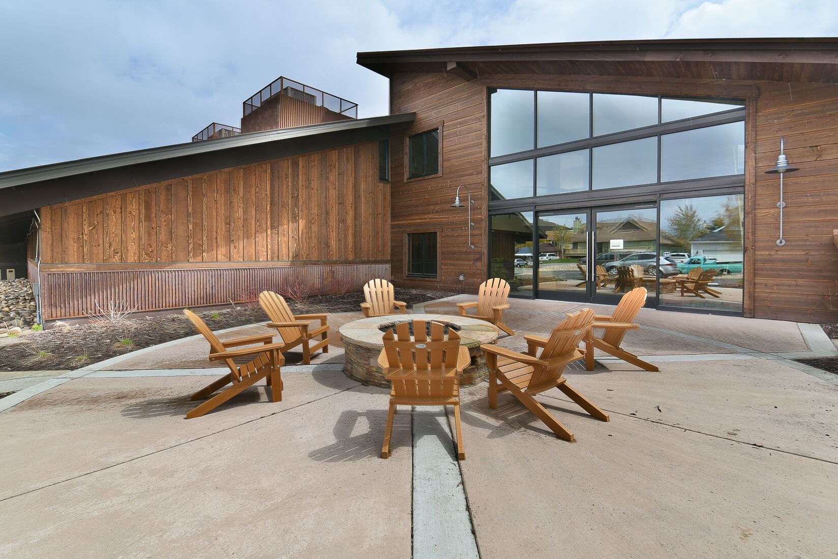 An outdoor seating area with Adirondack chairs and a fire pit, adjacent to a building with large windows and wooden siding.