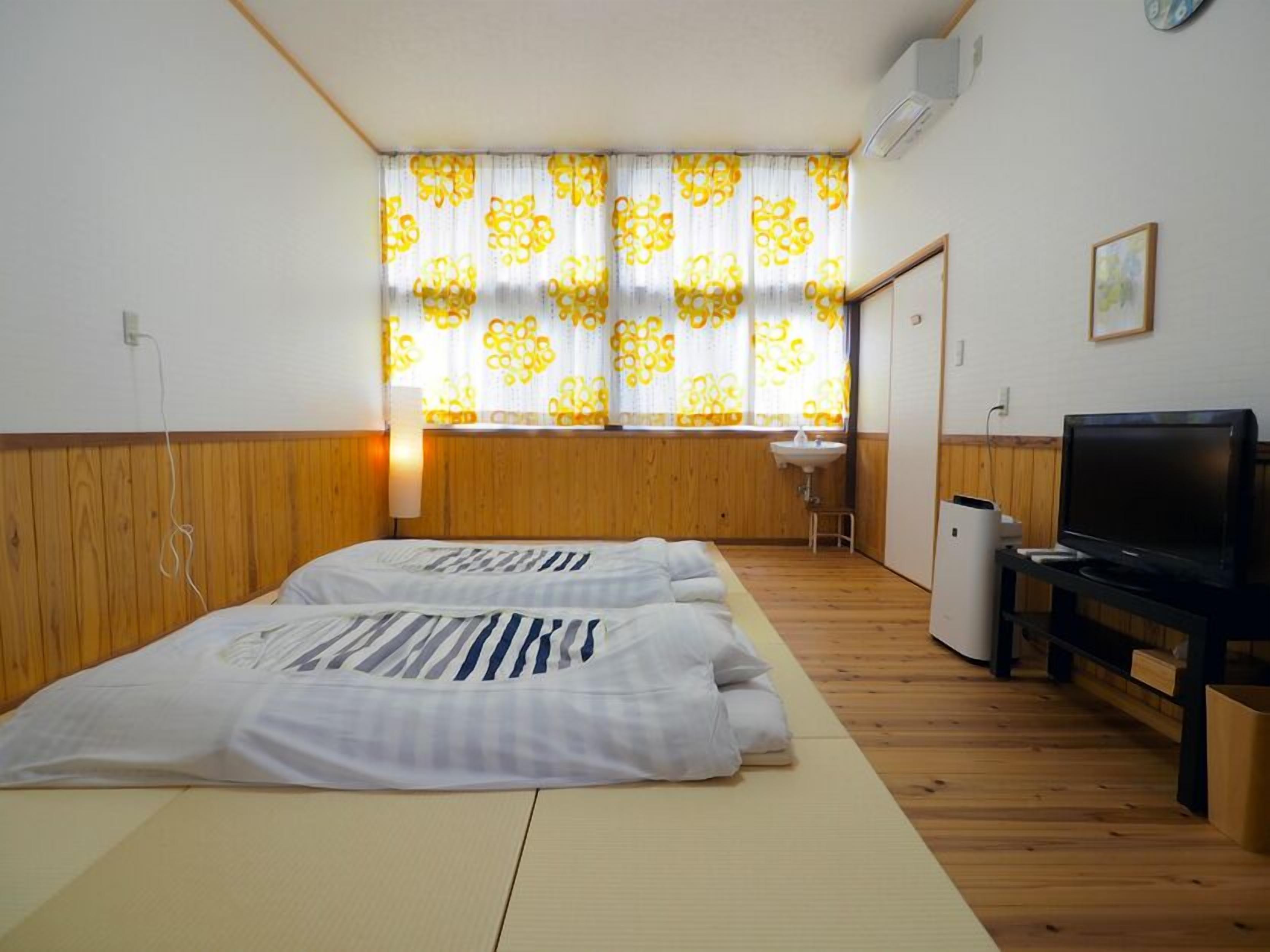 A room with two beds, a TV, a sink, and a window with patterned curtains.