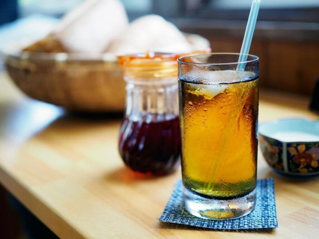 A glass of iced tea with a straw on a wooden table, accompanied by a small pitcher and a bowl of cookies.
