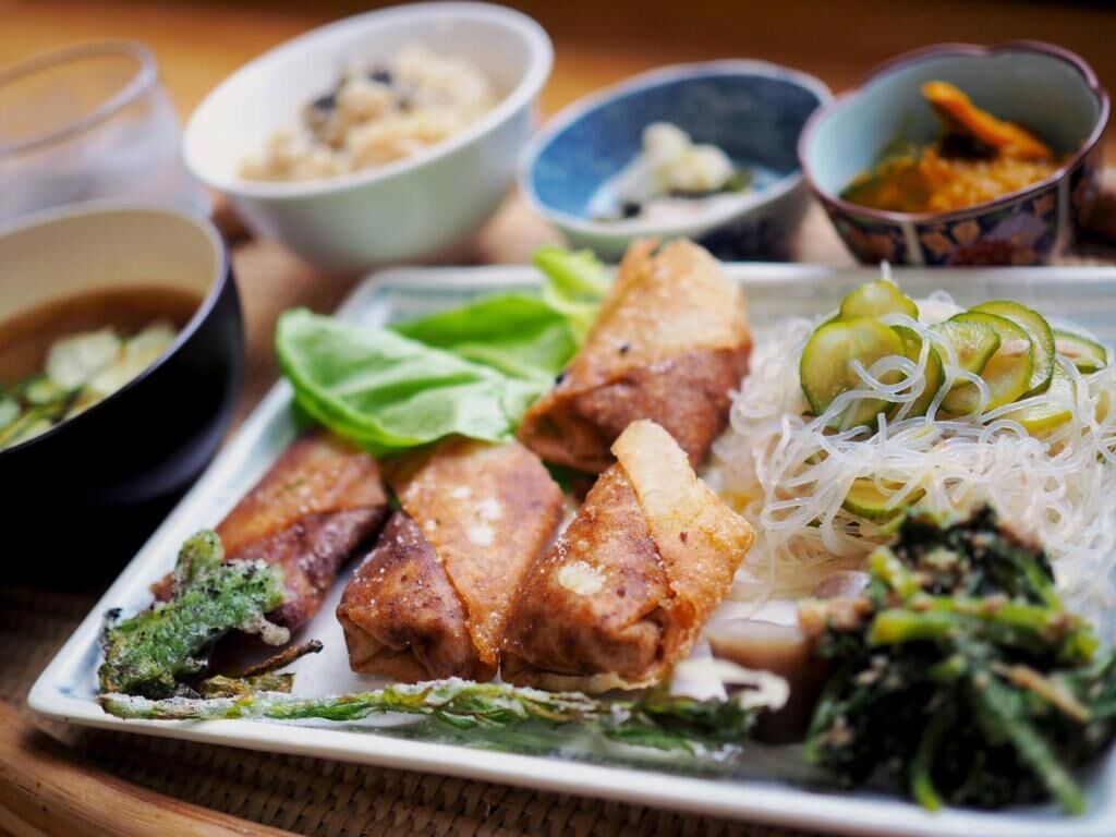 A plate of food with various dishes including fried items, noodles, and greens, accompanied by small bowls of sauces or condiments.