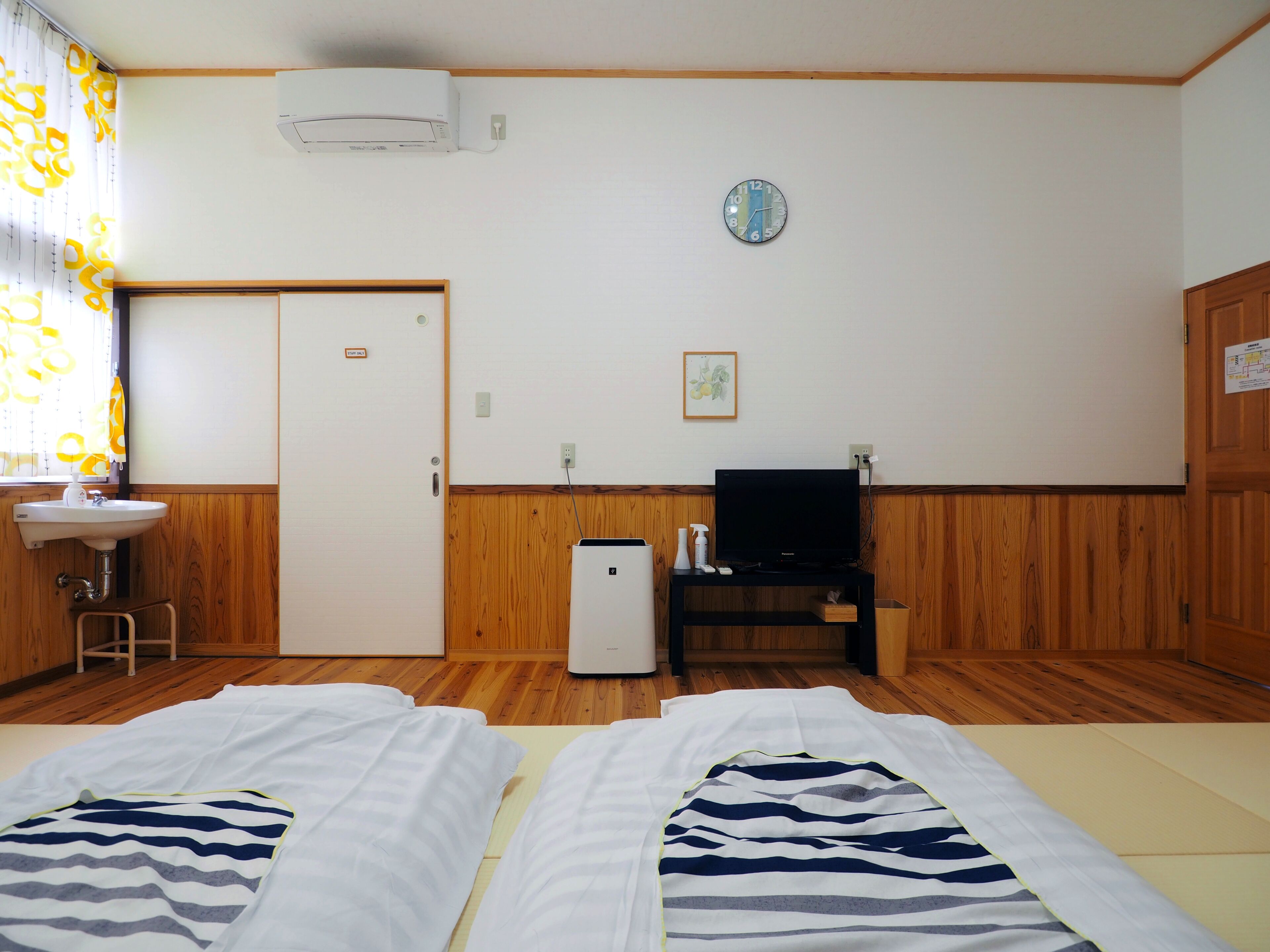 A room with two beds, a television, a small table, a sink, and a clock on the wall.