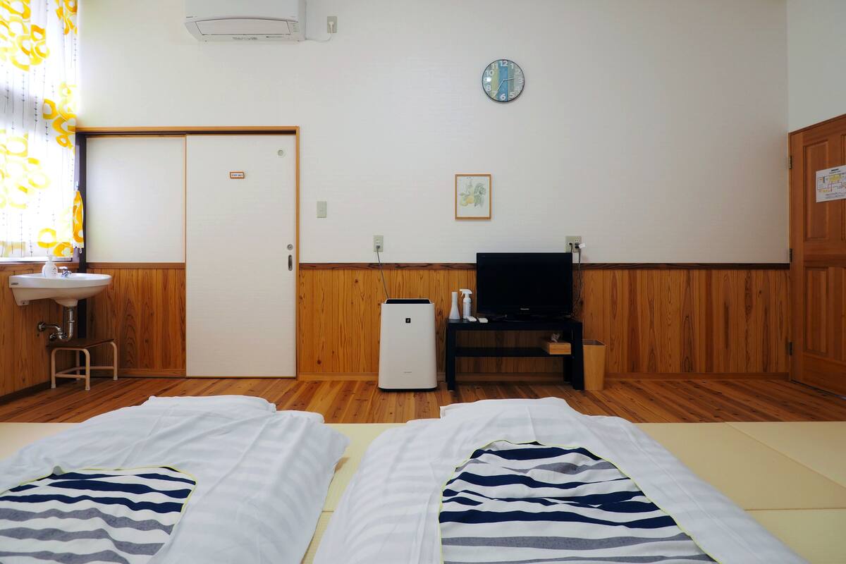 A room with two beds, a television, a small table, a sink, and a clock on the wall.