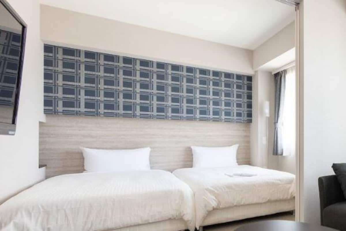 A hotel room with two beds, a sofa, a large window with curtains, and a wall-mounted TV.