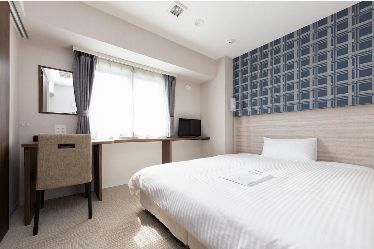 A hotel room with a bed, a desk, a chair, a TV, and a patterned wall.