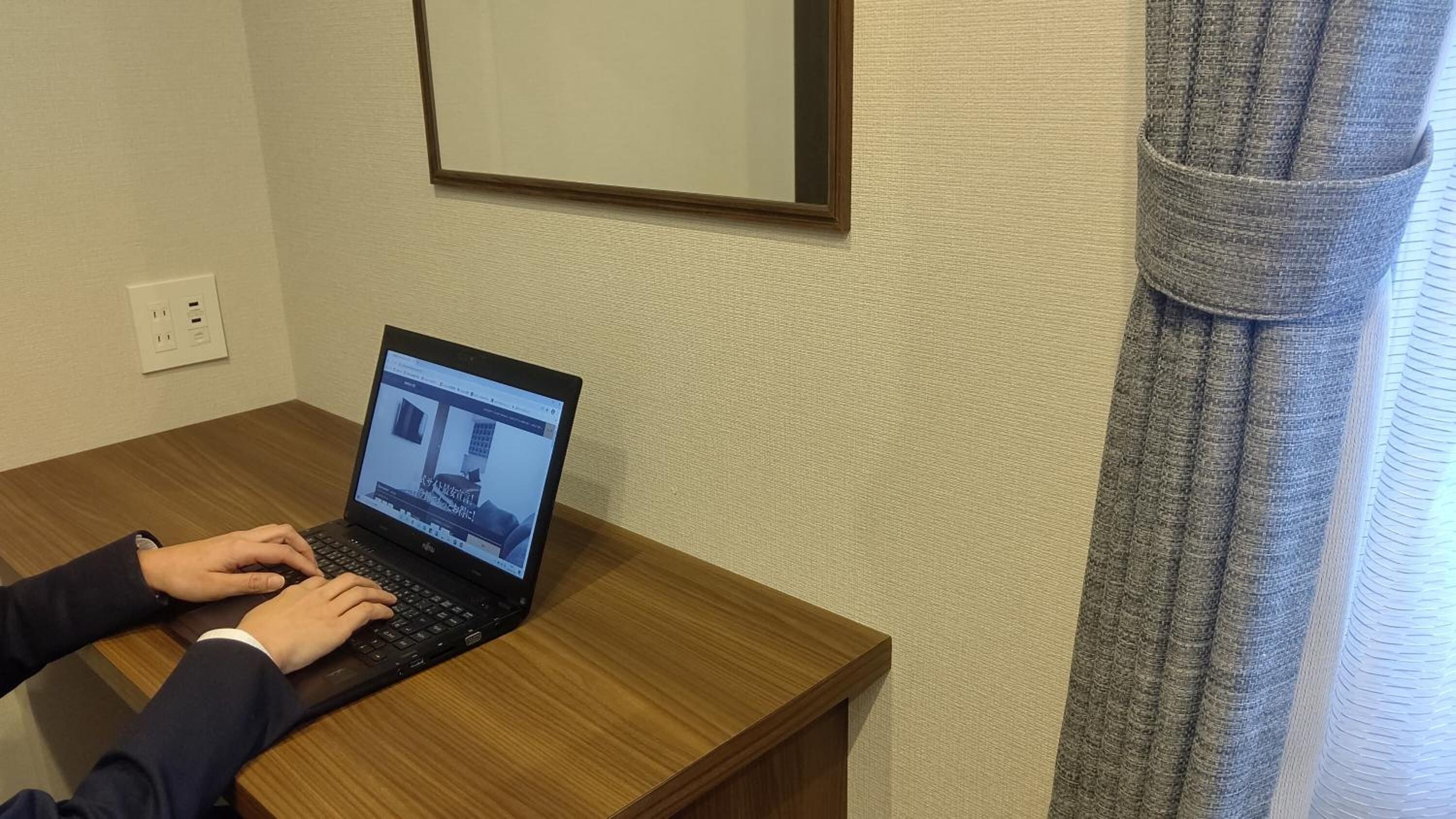 Una persona usando una computadora portátil sobre una mesa de madera en una habitación con pared lisa y una ventana con cortinas.
