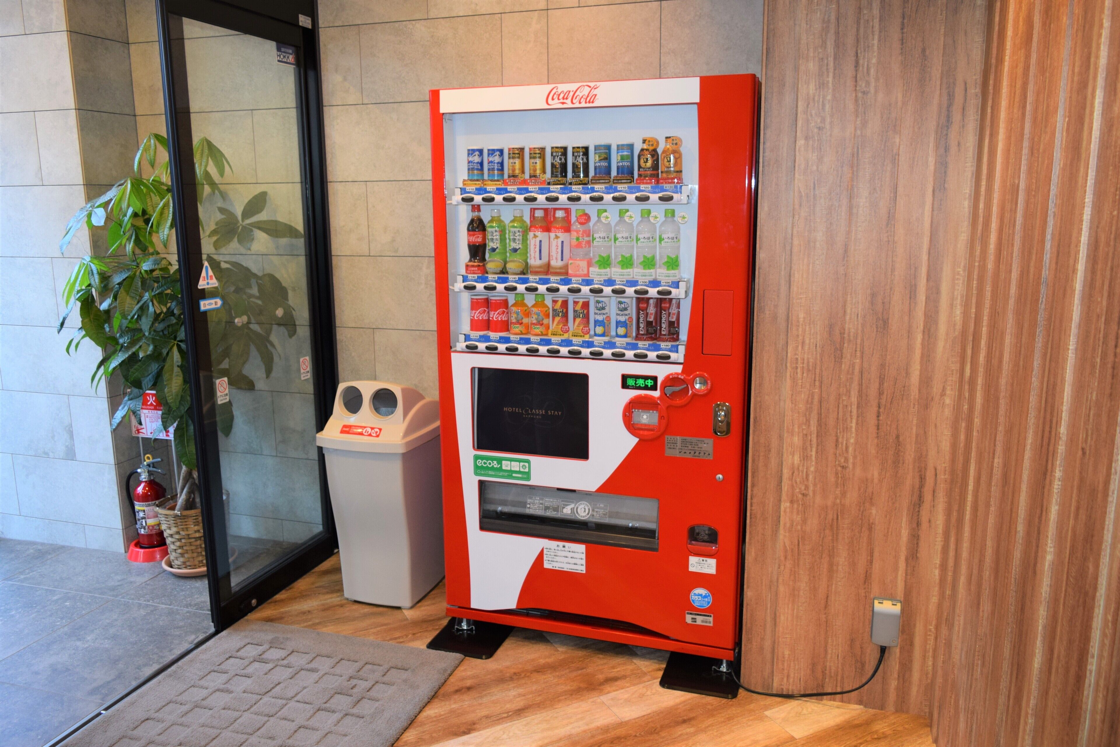 A Coca-Cola vending machine with various drinks, a fire extinguisher, and a trash bin.