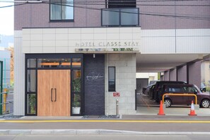 A hotel entrance with a wooden door, a black sign above the door, and a parking area with cars and orange traffic cones. - Hotel Classe Stay Sapporo (Sapporo)