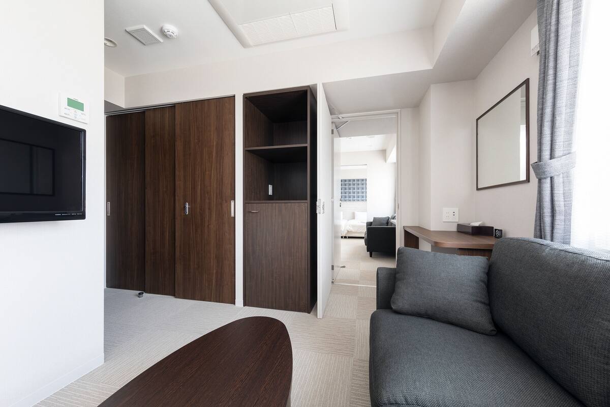 A modern hotel room with a grey sofa, a wooden wardrobe, a desk, and a large mirror.
