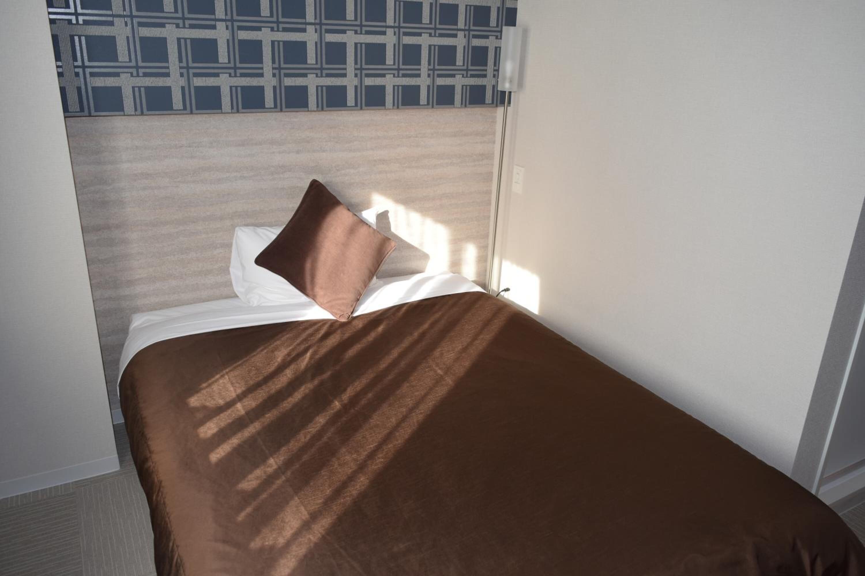Ein ordentlich bezogenes Bett mit einer braunen Tagesdecke und einem Kissen, vor einer Wand mit einem geometrischen Muster.