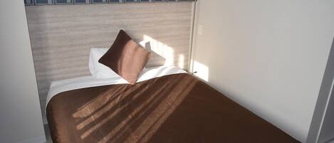 Ein ordentlich bezogenes Bett mit einer braunen Tagesdecke und einem Kissen, vor einer Wand mit einem geometrischen Muster.