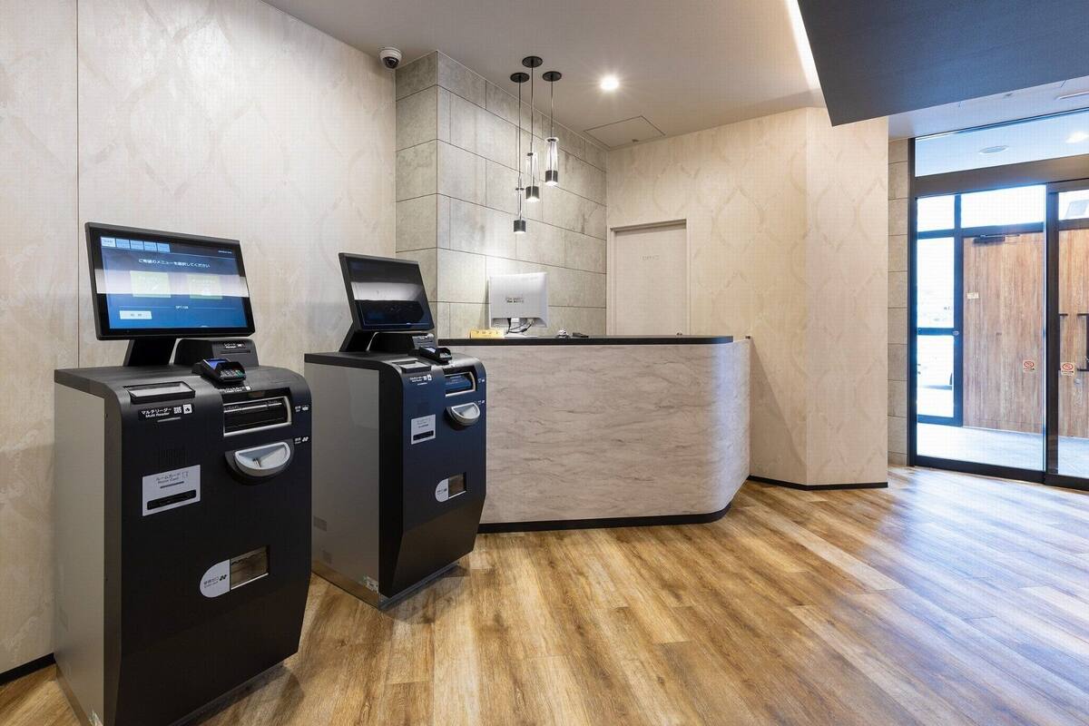 A reception area with a modern design, including a reception desk, two self-service kiosks, a security camera, and a wooden floor.
