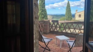 A balcony with two chairs and a small table, overlooking a rural landscape with stone buildings and tall trees. - Holiday apartment with pool and terrace (Asciano)