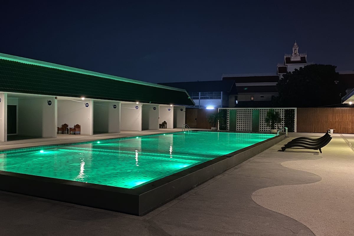 A swimming pool with a green-lit edge, a covered seating area with chairs, and a view of a building with a clock tower in the background.