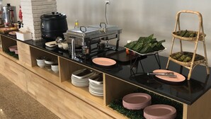 Montaje de bufé con diversos alimentos y utensilios, que incluye una máquina de café, un recipiente negro grande y una cesta de mimbre con elementos verdes.