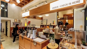 A cafe with a wooden counter, a coffee machine, and a sign that says "Order / Check In Here". - StayAt Bedever Bangkok Hotel (Bangkok)