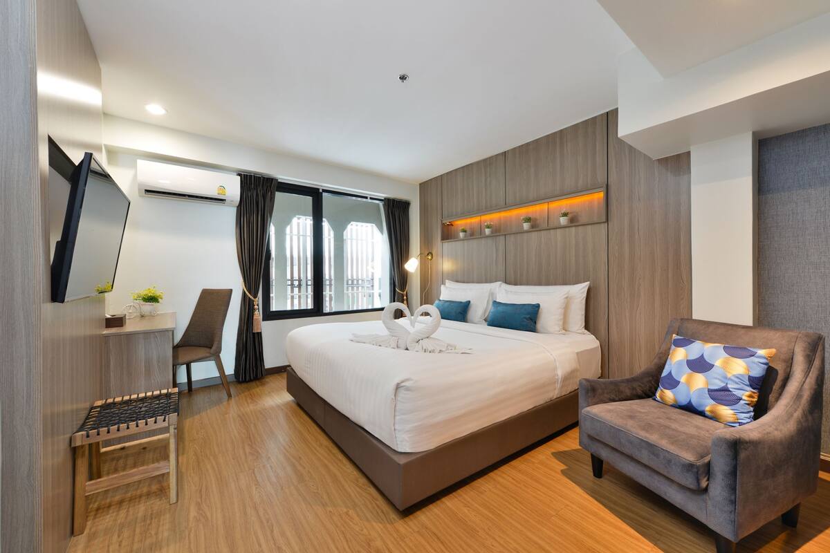 A modern hotel room with a large bed, a seating area, a desk, and a built-in air conditioning unit.