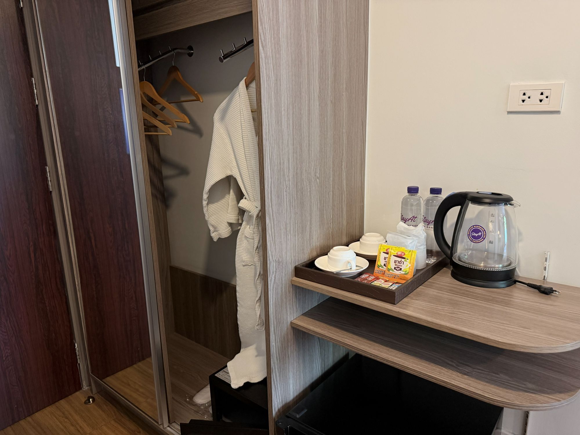 Habitación de hotel con suelo de madera, mampara de vidrio en la ducha, una toalla en un perchero, bandeja con artículos de aseo, tetera y toma de corriente en la pared.