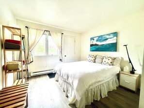 A bedroom with a bed, a nightstand, a lamp, a window with curtains, and a painting on the wall.
