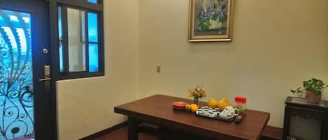 Una habitación con una mesa de madera, un cuadro enmarcado y varios objetos sobre la mesa, entre ellos frutas, un florero y una pequeña planta.