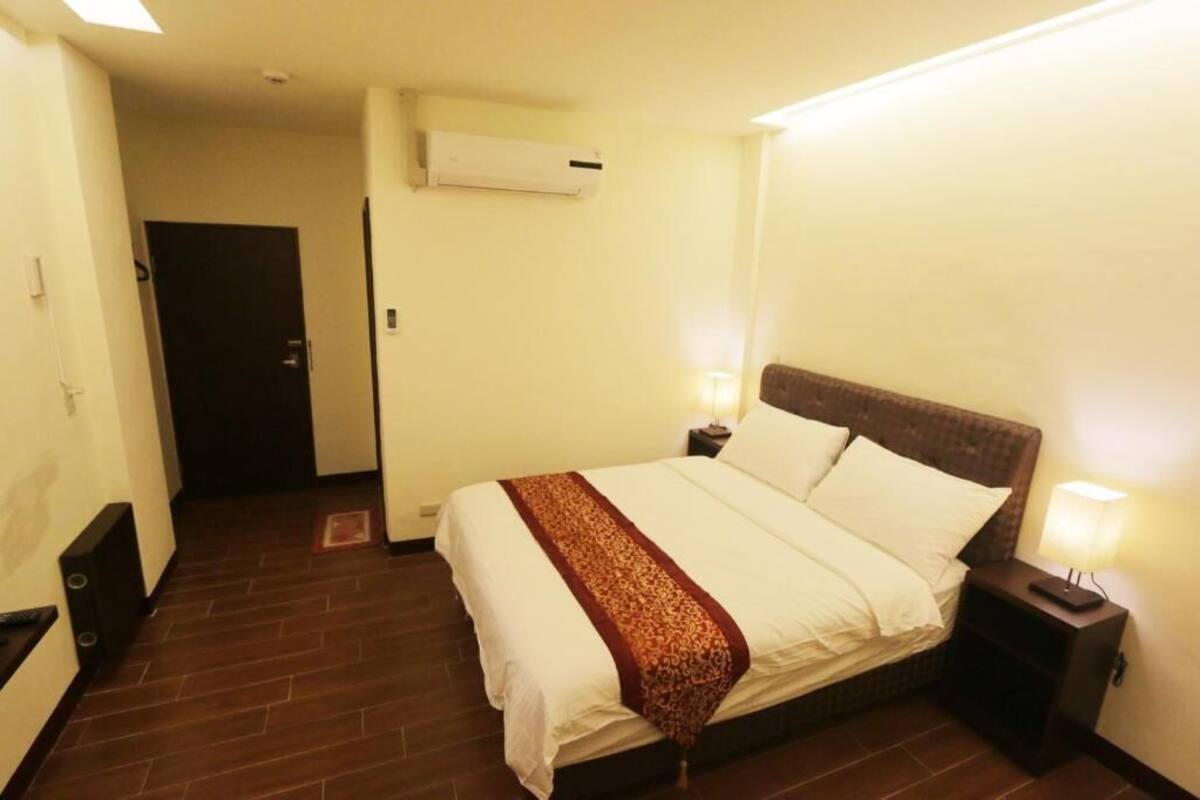 A hotel room with a bed, bedside lamps, an air conditioning unit, and a door.