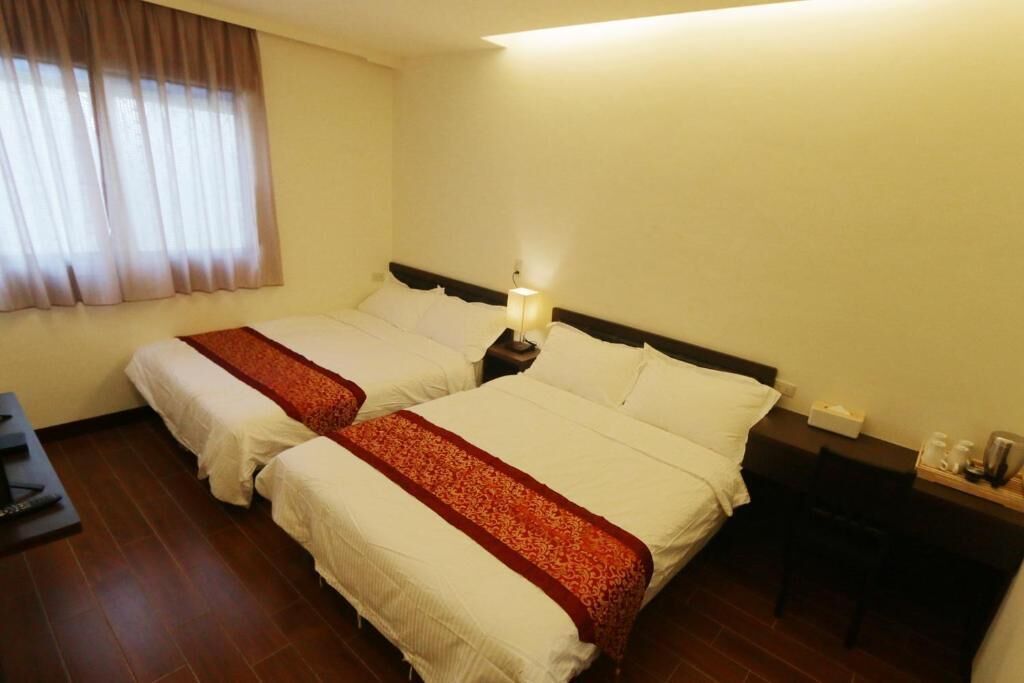 A hotel room with two beds, a desk, a chair, and a nightstand with a lamp.