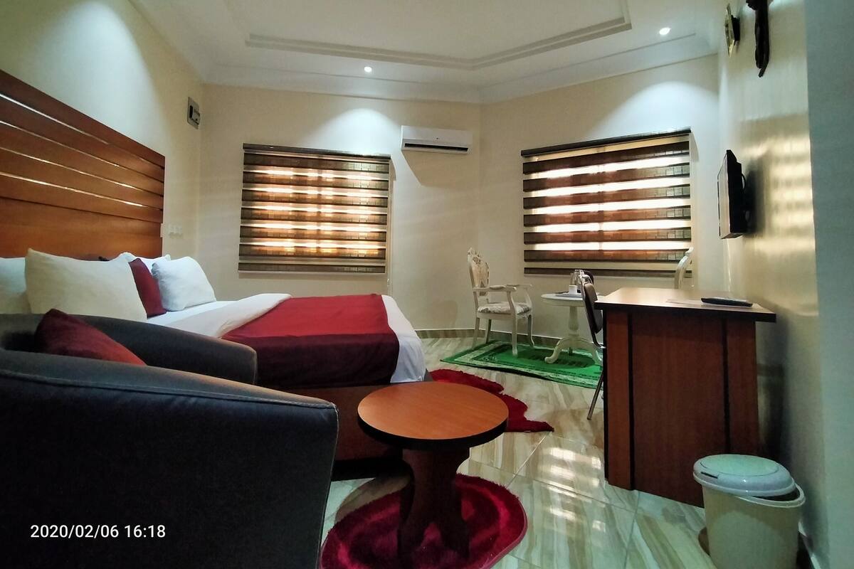 A hotel room with a bed, a sofa, a small table, a chair, a desk, and a trash bin.