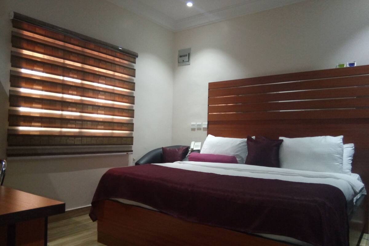 A hotel room with a wooden headboard, a bed with white and purple bedding, a desk with a chair, and a window with blinds.