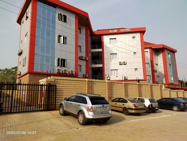 A multi-story building with the sign "ALIM ROYAL" on the facade, featuring a modern architectural design with red and white colors, multiple air conditioning units, and a parking area with several cars.