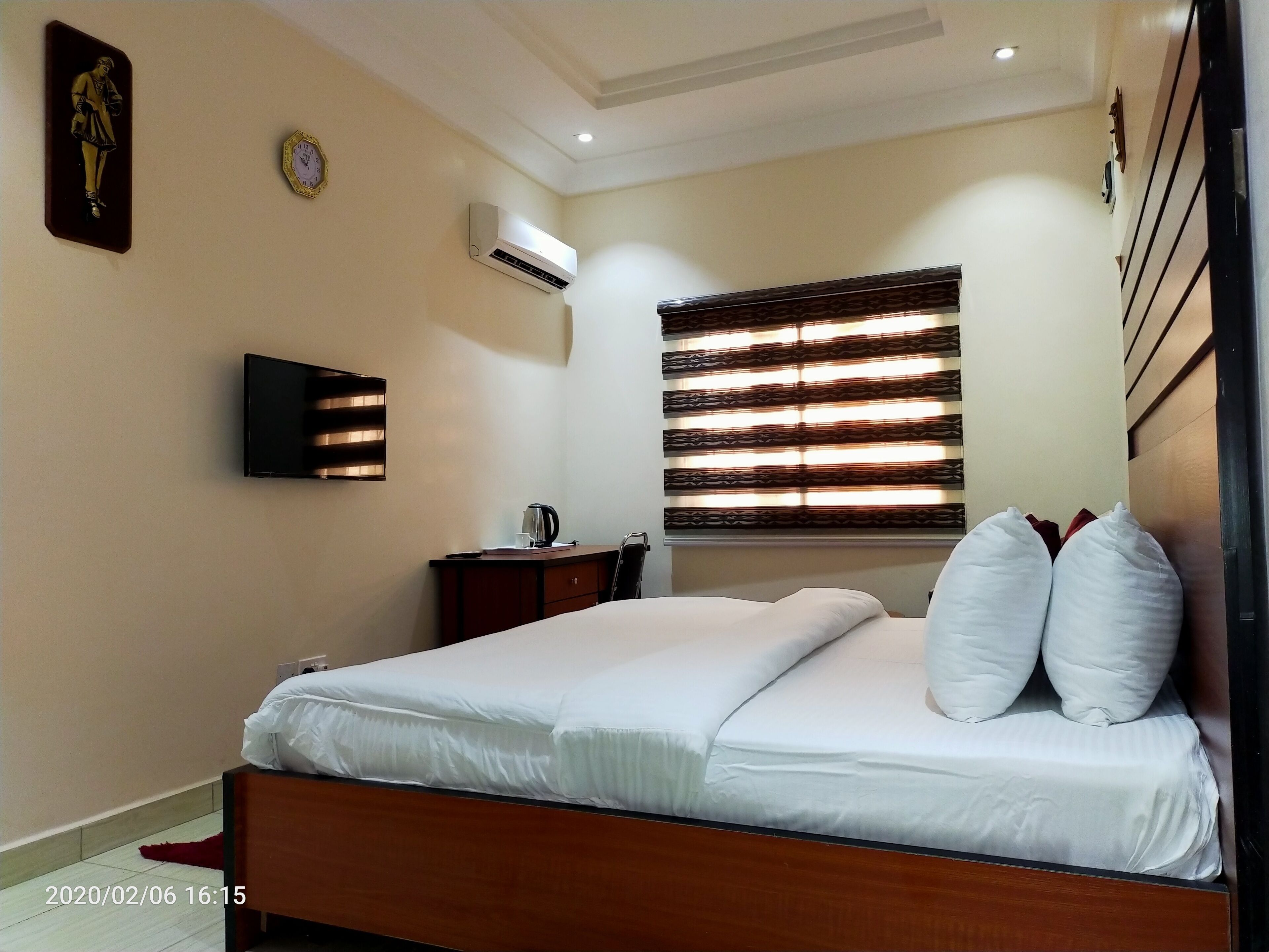 A neatly made bed with white linens and pillows, a bedside table with a coffee maker, a wall-mounted TV, and a window with blinds.