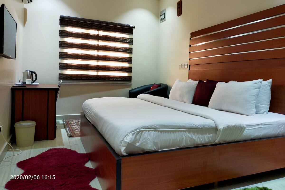A modern bedroom with a wooden bed, a bedside table with a kettle, and a window with blinds.
