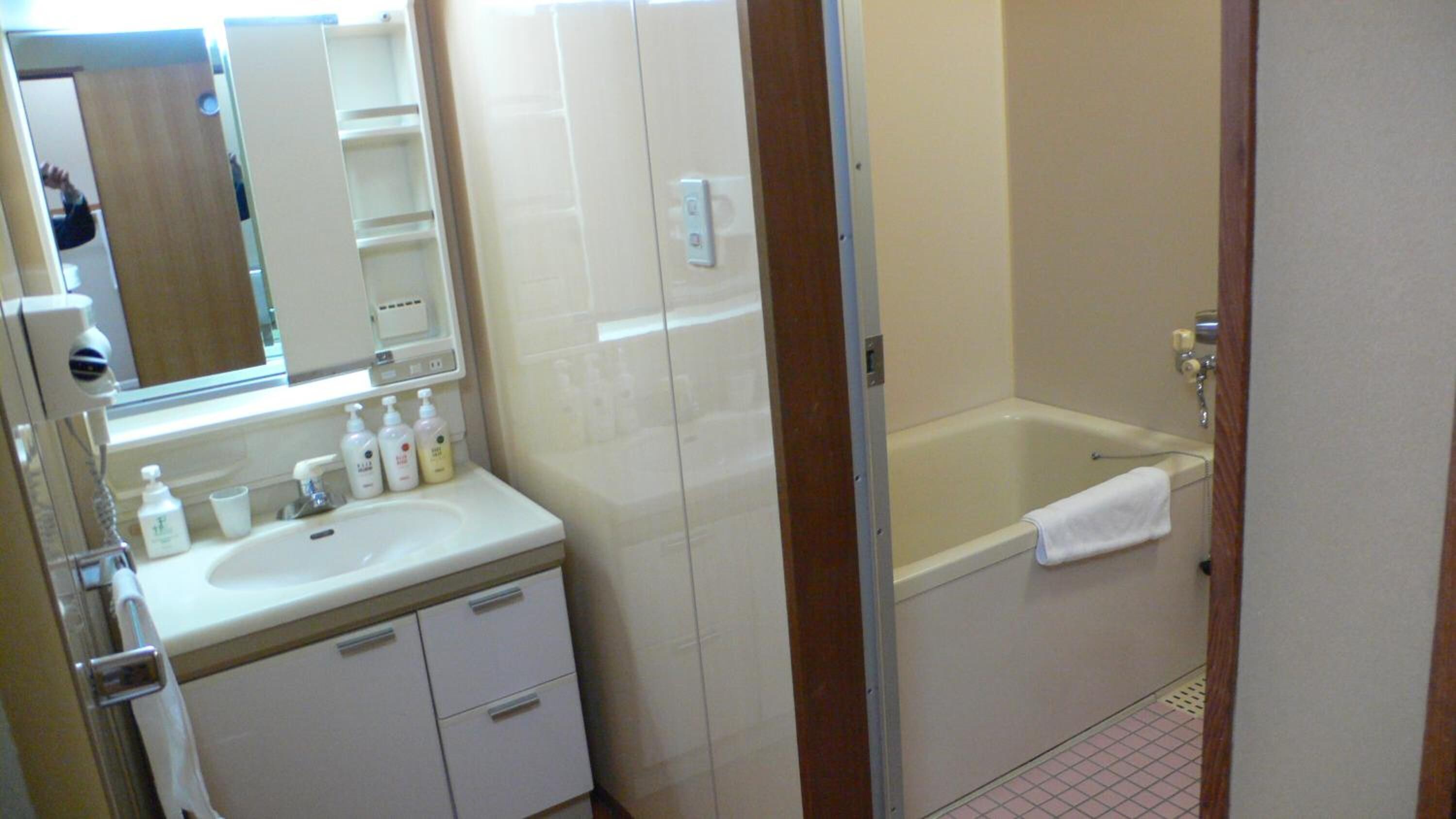 A bathroom with a white sink, mirror, and toiletries. There is a bathtub with a towel.