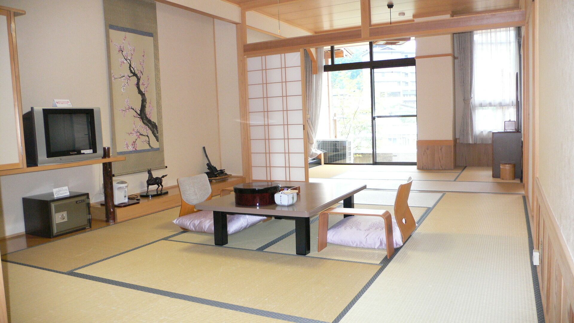 A traditional Japanese-style room with tatami flooring, a low table with cushions, a television on a shelf, and sliding doors.
