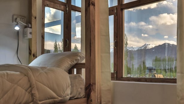 A bed with a quilted blanket in a room with large windows, wooden beams, and a view of mountains. - Ree Hostel (Leh)