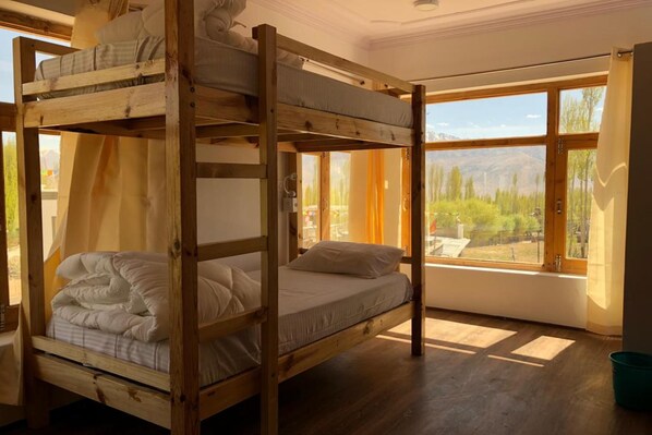 A bunk bed room with wooden frames, a window with curtains, and a view of greenery outside. - Ree Hostel (Leh)