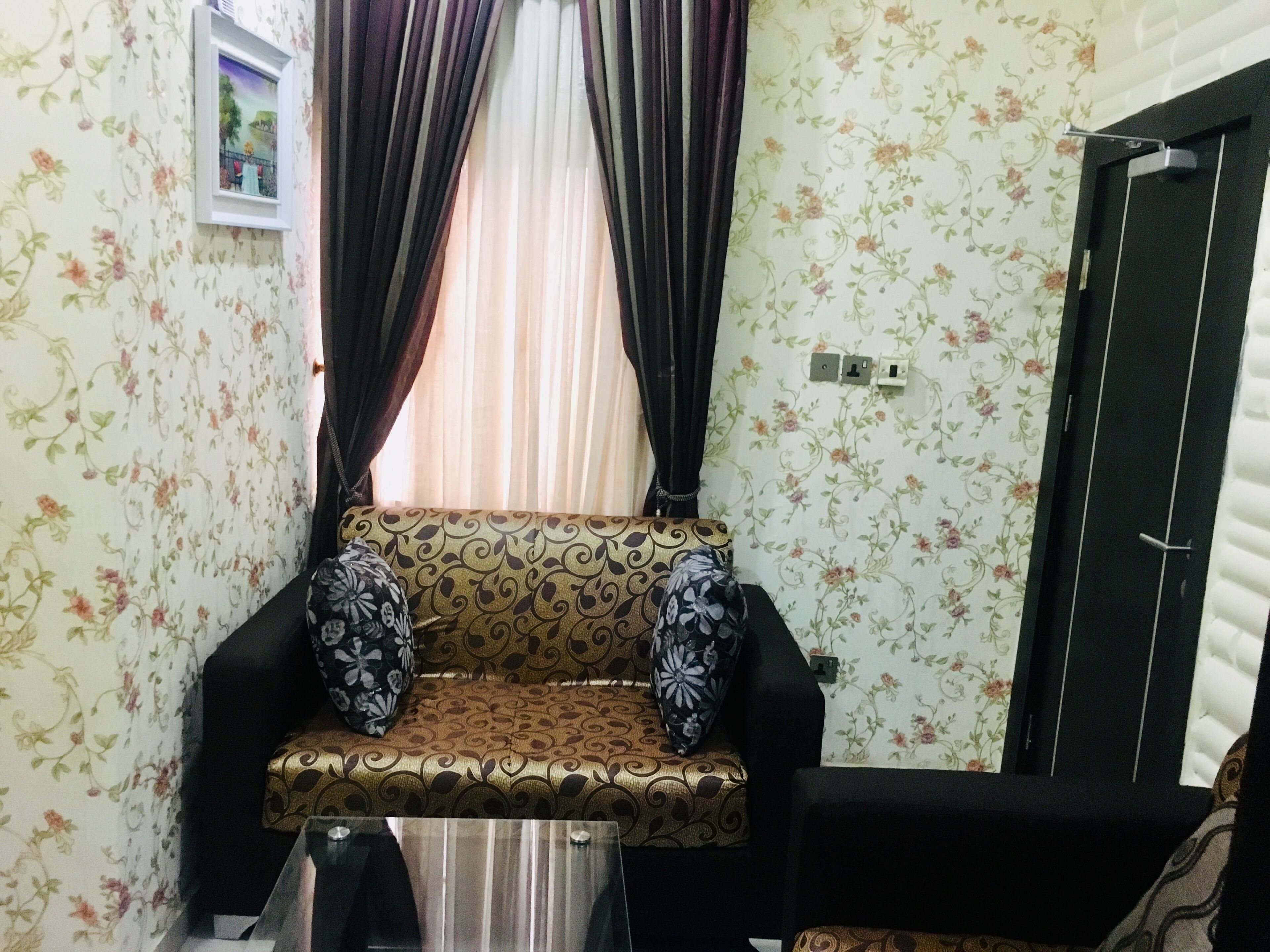 A room with floral wallpaper, a sofa with patterned cushions, a window with curtains, and a door.