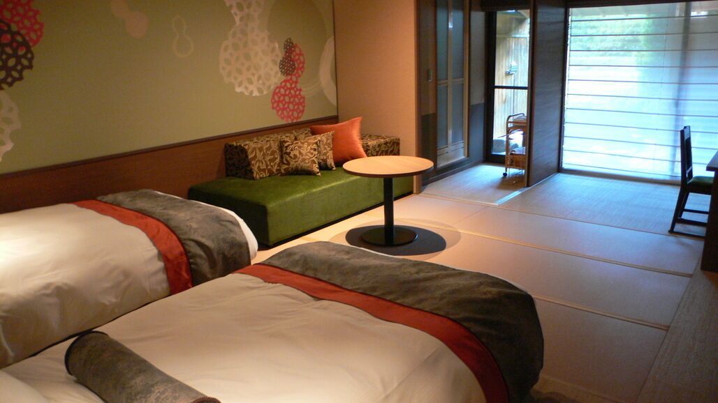 Une chambre d’hôtel avec deux lits, un canapé vert, une petite table ronde et une vue sur l’extérieur grâce à de grandes fenêtres.