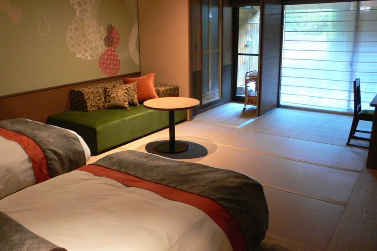 Une chambre d’hôtel avec deux lits, un canapé vert, une petite table ronde et une vue sur l’extérieur grâce à de grandes fenêtres.