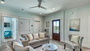 A spacious living room with a large grey sofa, a patterned armchair, and a round coffee table. There is a ceiling fan, a door with a view, and a mounted artwork on the wall. - SEA YOU SOON: Gorgeous Home in Gated Neighborhood w/ Private Pool & Easy Beach Access! (Miramar Beach)