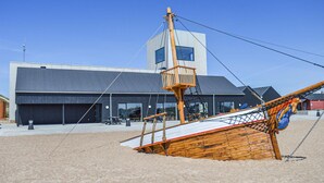 A wooden shipwreck on a sandy beach with a modern building in the background.