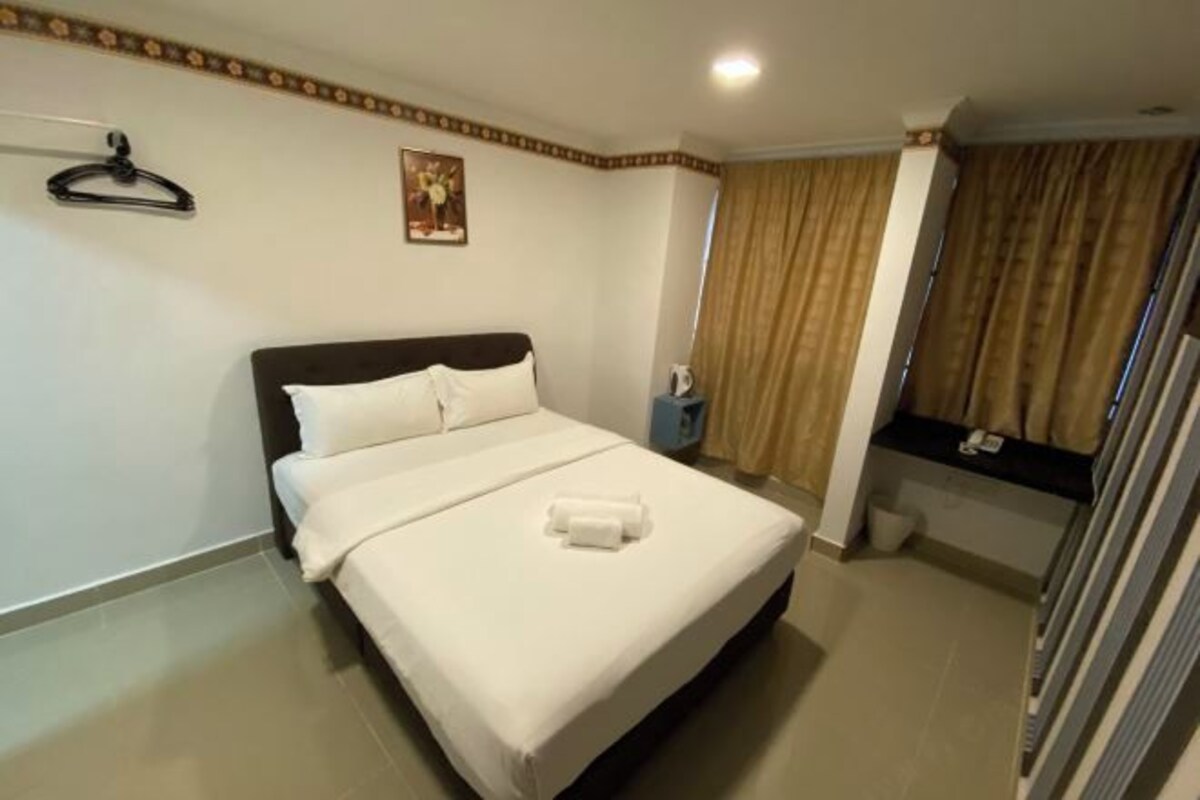 A neatly made bed with white linens and a rolled towel, a small bedside table with a lamp, a framed picture on the wall, and a window with curtains.
