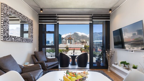 A modern living room with a large window, a dining table with fruit, and a city view. - The Docklands 509 by CTHA (Cape Town)