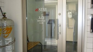 Entrada de hotel con puerta de vidrio, un banco en el interior y dos cilindros de gas afuera.