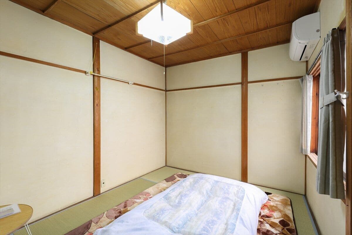 A small, traditional Japanese-style room with tatami flooring, a bed with a floral-patterned bedspread, a wooden ceiling, and a window with curtains.