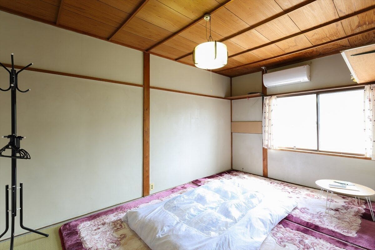 A room with a bed, a small table, a coat rack, and a window with curtains.