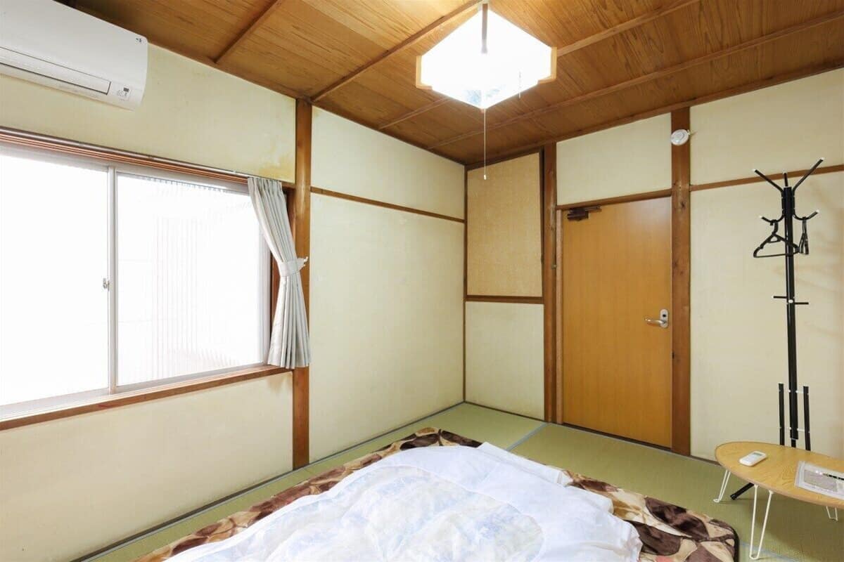 A traditional Japanese room with tatami flooring, a bed with a white blanket, a wooden door, and a window with curtains.