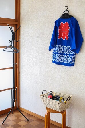 Une pièce avec un plancher en bois, un porte-manteau fixé au mur, un vêtement suspendu aux motifs rouges et blancs, et un panier en osier contenant des objets posés sur un tabouret.