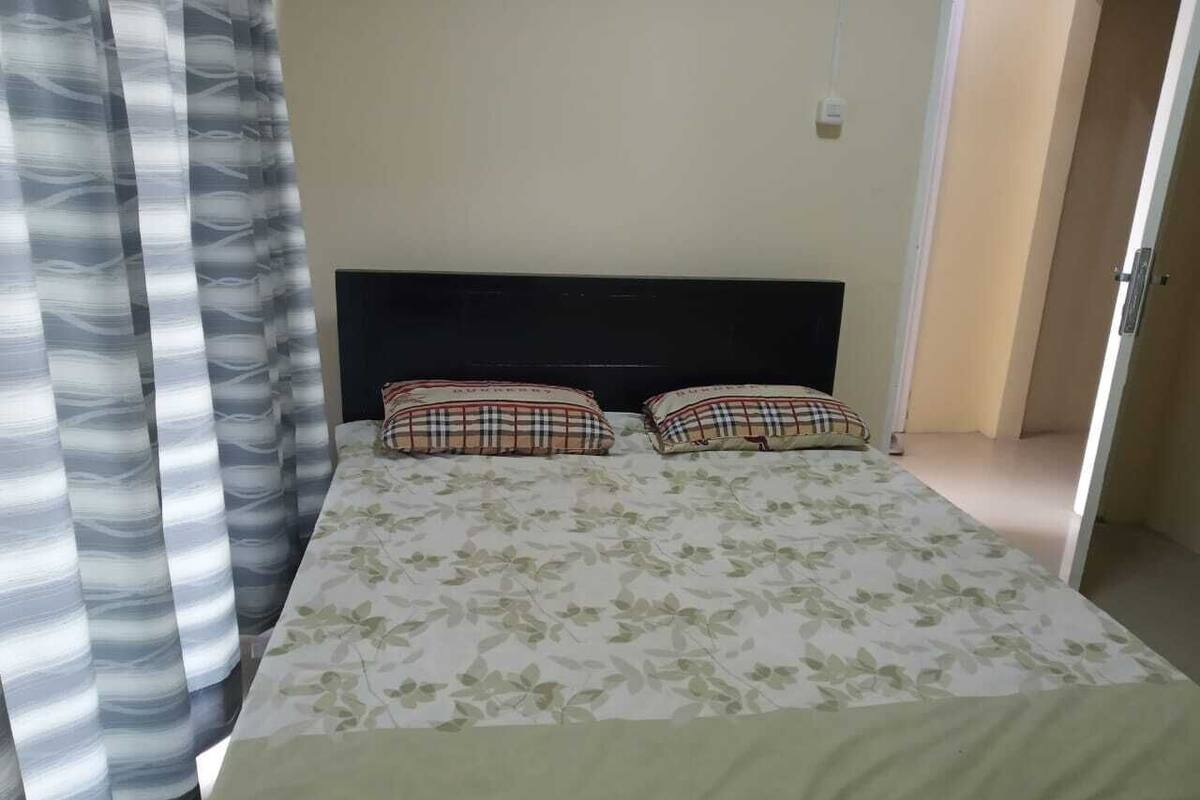 Un lit avec une couvre-lit à motifs floraux et des coussins à carreaux, une tête de lit sombre et une fenêtre avec des rideaux à rayures.